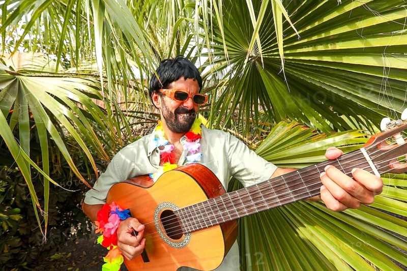A cheerful man wearing sunglasses, a wig and a colorful flower necklace plays a guitar surrounded by lush palm leaves, depicting a carefree atmosphere of tropical vacation and friendly fun