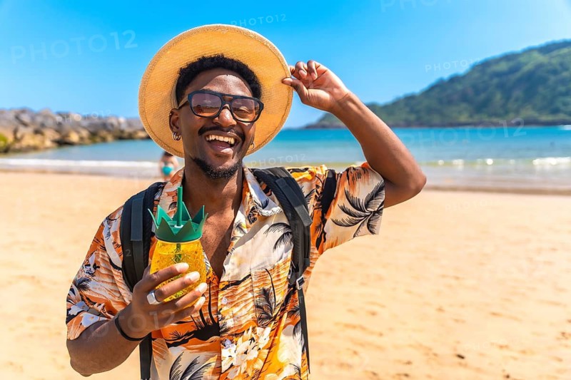 Young black man wearing a straw hat and sunglasses, savoring a refreshing pineapple cocktail while relaxing on a sunny beach