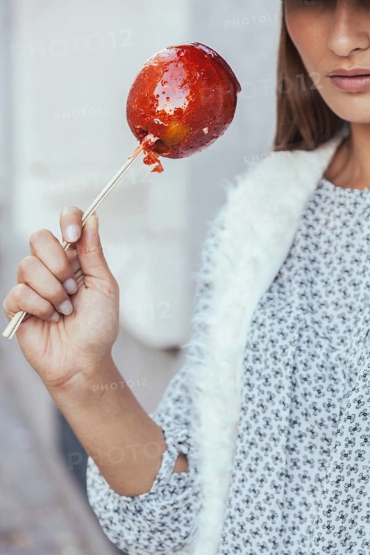 Close-up of a woman holding a glossy red candy apple on a stick. She's dressed in casual autumn attire, with focus on the sugary treat, capturing a cozy fall vibe