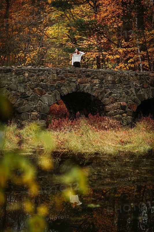 A woman stands on a stone bridge overlooking a tranquil pond surrounded by vivid autumn foliage in Quebec, Canada. The scene captures the serene beauty of fall