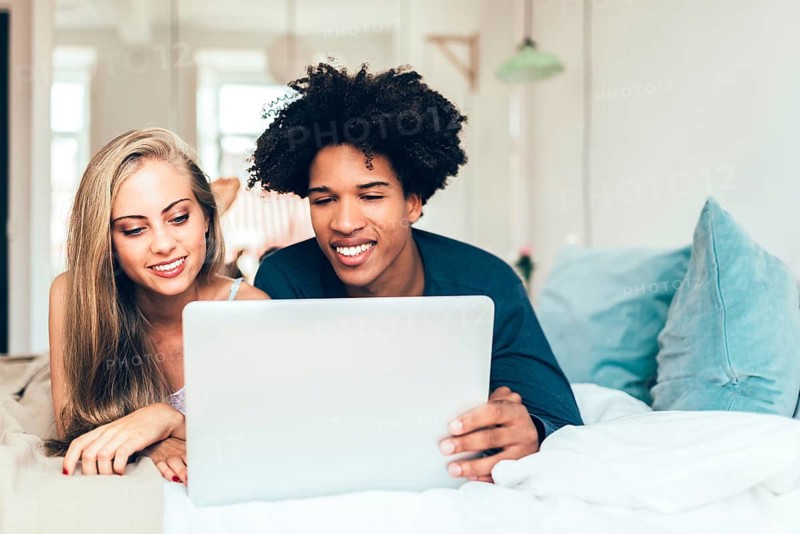 A cheerful couple is lounging on a bed, focused on a laptop screen. The cozy setting and relaxed ambiance highlight leisure and technology in a modern home environment