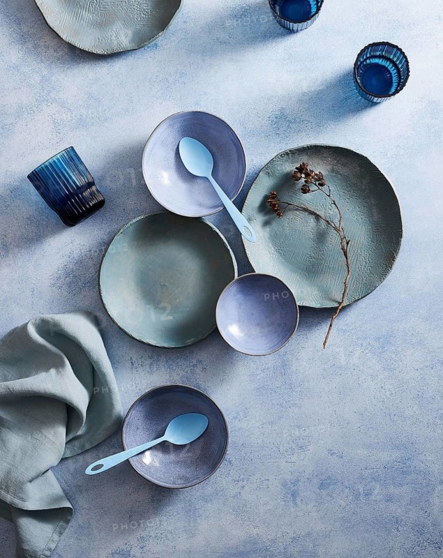 Top view of a stylish ceramic tableware set, featuring pastel-colored plates, bowls, and cups on a textured background, complemented by a blue glass and delicate floral accents