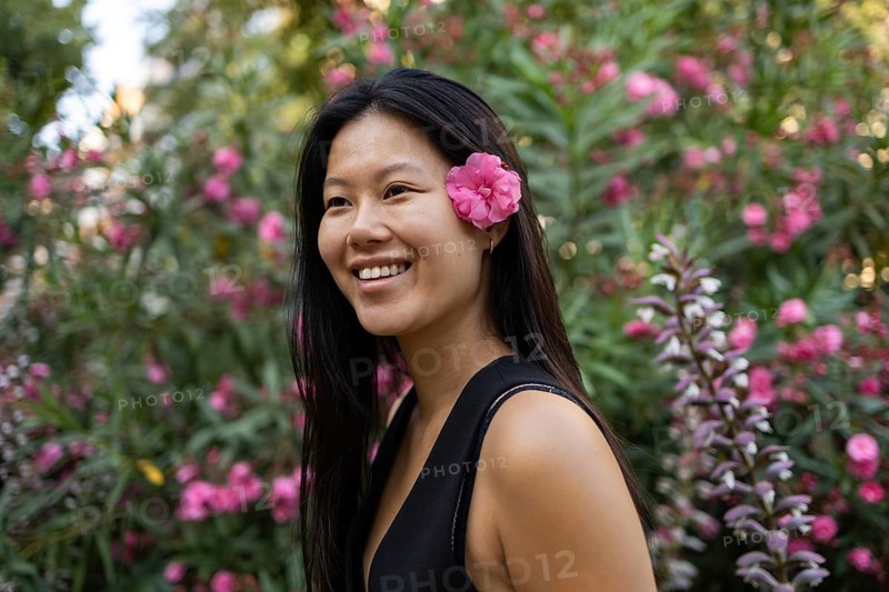 A Chinese woman with a pink flower in her hair smiles joyfully during a sunny summer day in the city. Lush greenery and pink blooms create a vibrant backdrop