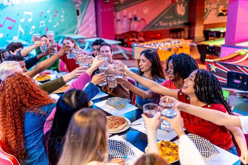 Cheerful young multiethnic friends raising glasses for a toast, enjoying food and drinks at a bowling alley restaurant