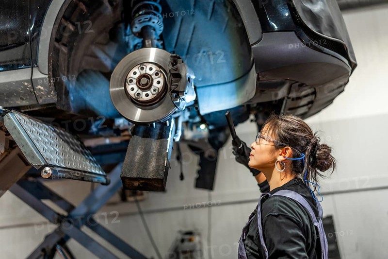 Female mechanic using flashlight, inspecting brakes of lifted car in a professional workshop, ensuring safety and performance