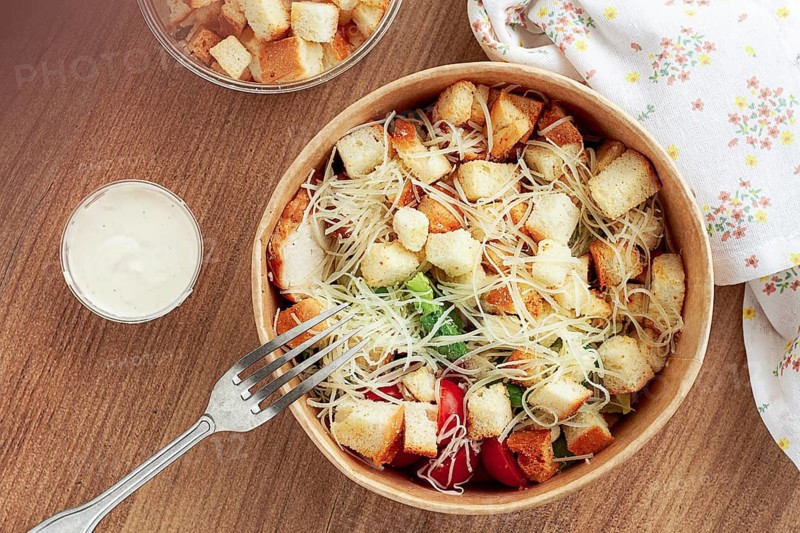 Caesar salad in a paper container, take it on the way, with sauce and croutons, on the table, snack, natural light, no people