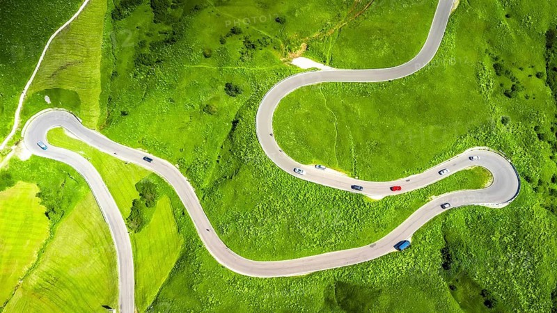 Winding road seen from above with cars driving on a green meadow in the italian dolomites mountains