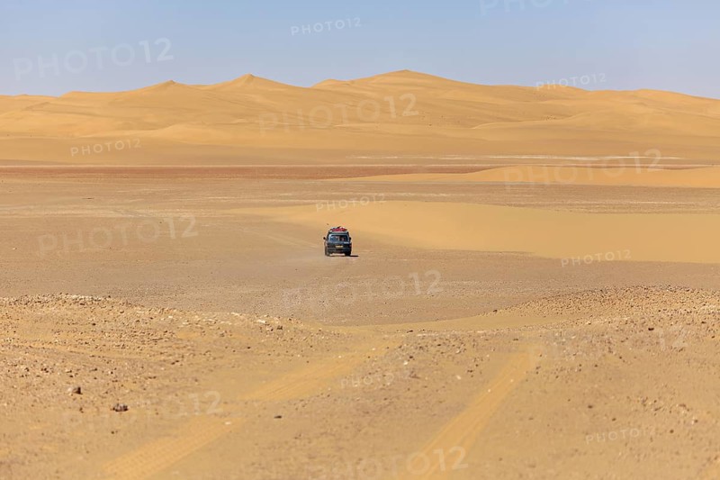 A car drives alone in a vast desert landscape with sand-coloured dunes and a clear sky, Off-road driving through the Sahara in Algeria
