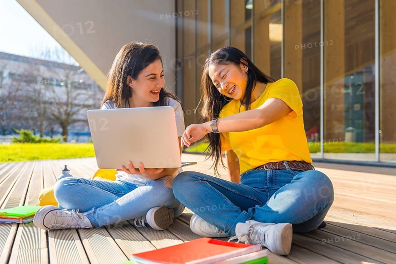 Two female multi-ethnic students sitting on the ground in the campus using laptop