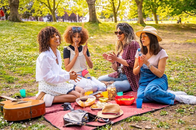 Four diverse female friends clapping hands, having fun during a picnic on a sunny day in a park, surrounded by food, drinks, and a guitar