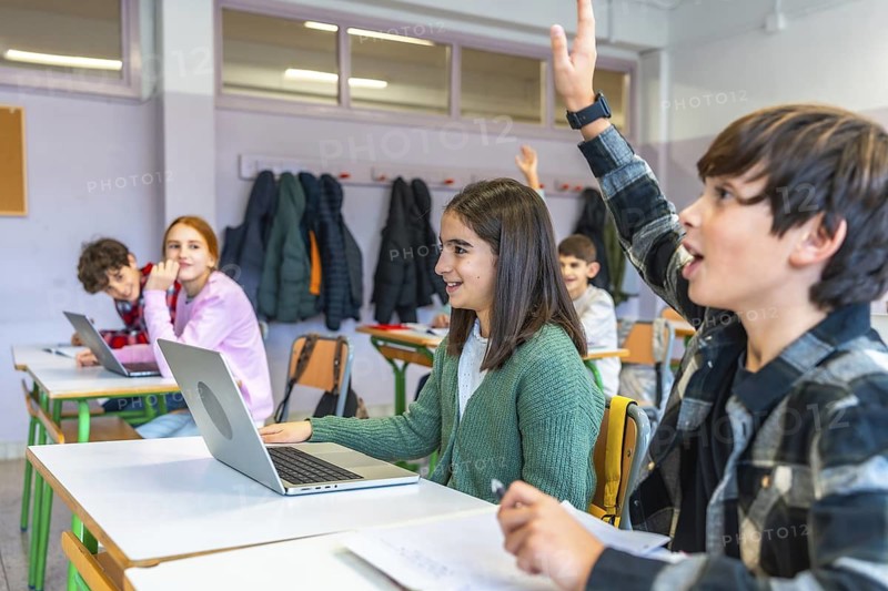Elementary school students raising hands and using laptops in an interactive classroom, engaging in modern educational activities