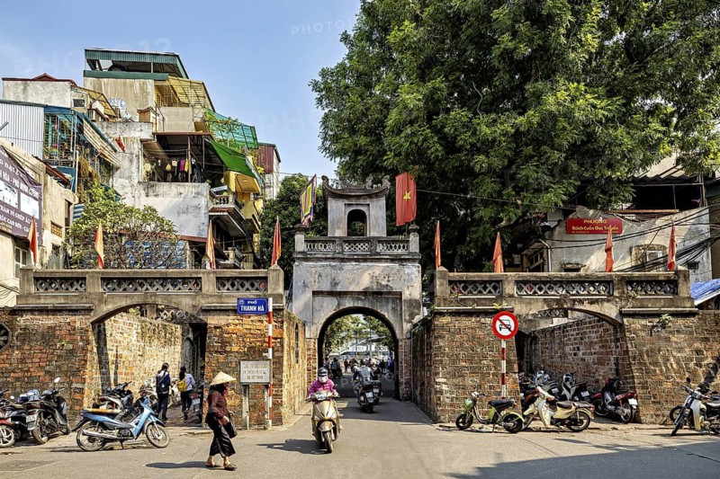 City gate in a busy street with motorbikes and passers-by, surrounded by historic buildings and trees on a sunny day, Historic city gate in the old quarter of Hanoi Vietnam