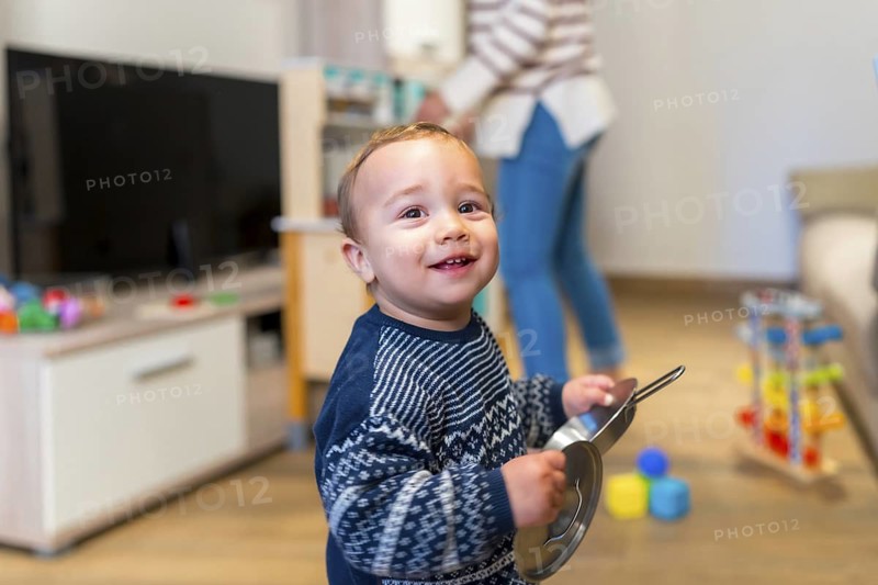 Smiling toddler playing with kitchenware in the living room while mother is tidying up in the background, concept of happy family life