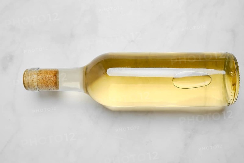 A glass bottle with white wine on a smooth light surface. The bottle has a cork top and shows the liquid inside, which appears clear and bright. Light reflects off the glass