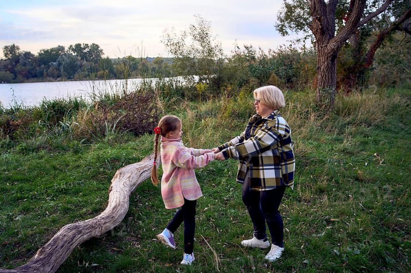 A mother and daughter enjoy a playful moment by the riverside, surrounded by lush greenery and trees. The girl is happily swinging, holding hands with her grandmother