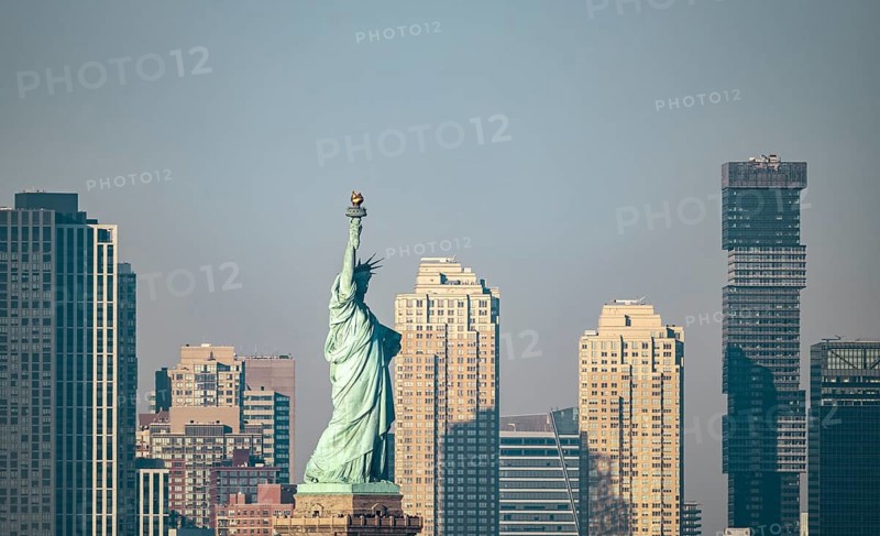 A clear view of the Statue of Liberty standing prominent against the backdrop of New York City's urban buildings, captured during the day under a soft light and hazy sky
