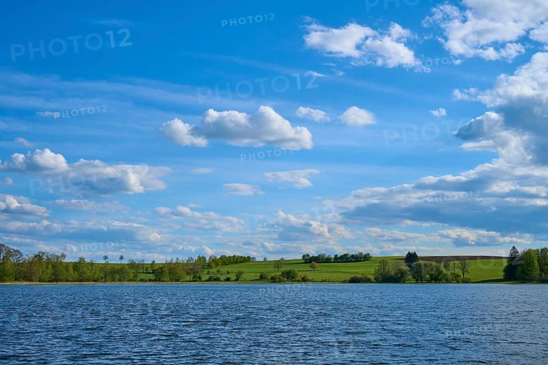 Ober Moosersee surrounded by green fields and trees under a partly cloudy blue sky, spring, Freiensteinau, Vogelsberg, Hesse, Germany