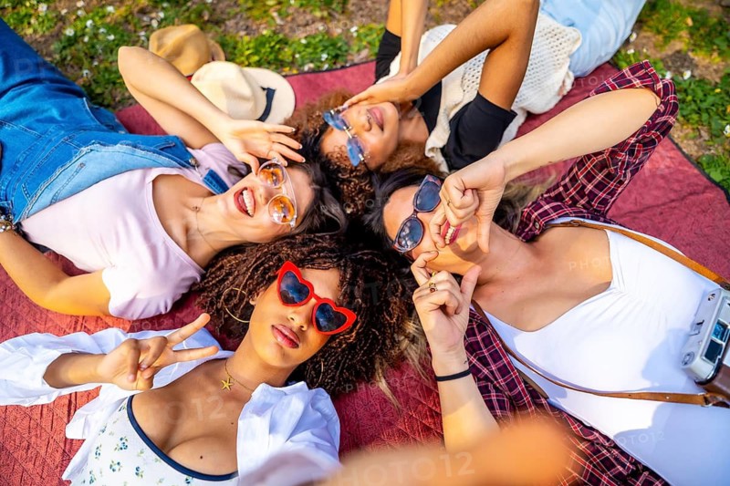 Four cheerful young women wearing sunglasses are lying on a blanket in a park, taking a selfie and making funny faces