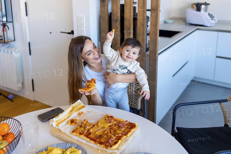 Happy mother holding her baby while eating pizza in a bright, modern kitchen, creating a heartwarming family moment
