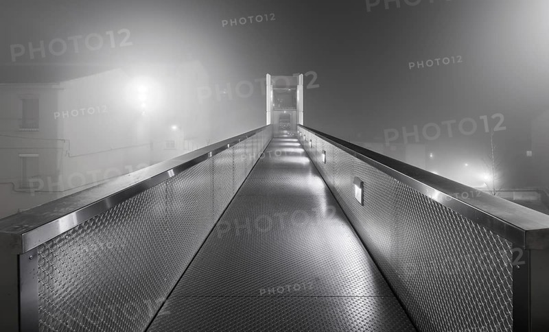 A dimly lit urban elevator walkway stretches into the misty fog, creating a mysterious and atmospheric scene at night