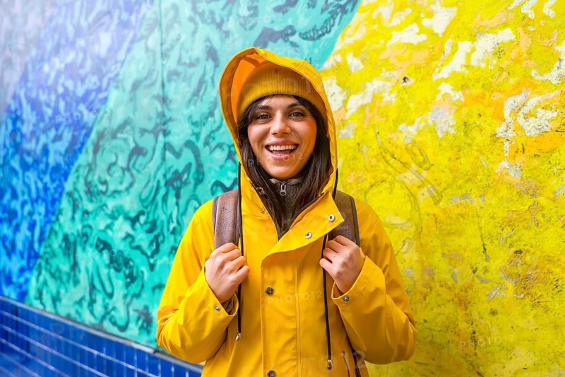 Young woman in a yellow raincoat and knit hat smiling brightly against a vibrant blue and yellow street art mural on san franciscos 16th avenue tiled steps, enjoying urban exploration