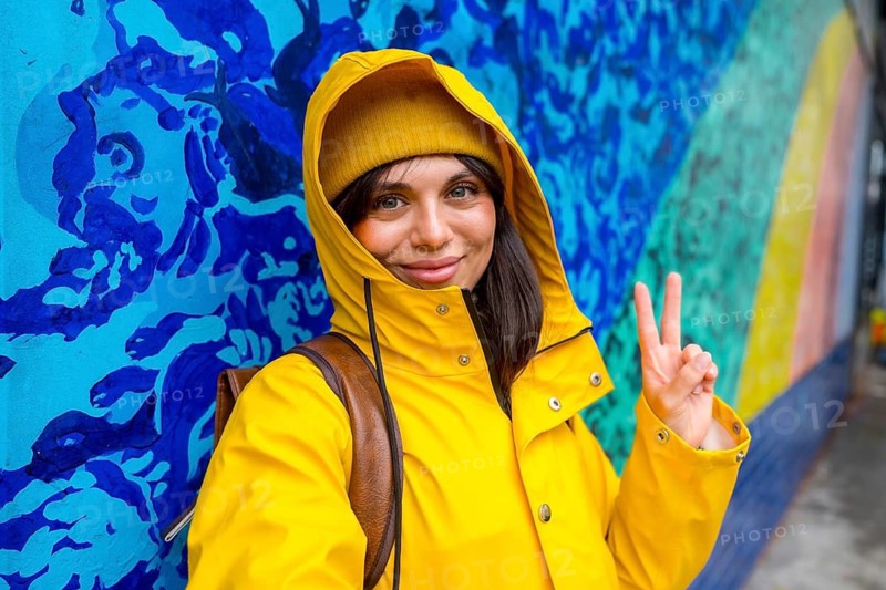 Young woman smiling, wearing a yellow raincoat and beanie, showing a peace sign against a vibrant blue and rainbow mural on a city street, expressing happiness and good vibes