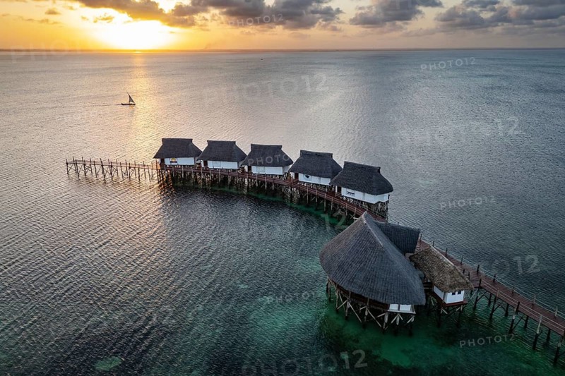Aerial view of luxury overwater bungalows with thatched roofs and a wooden pier surrounded by emerald ocean and white sand beach in Zanzibar
