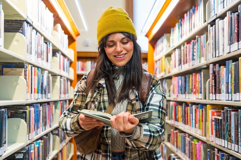 Young woman standing between rows of diverse bookshelves, focused on reading a book in her hands while smiling, enjoying learning and studying in a vibrant san francisco library setting