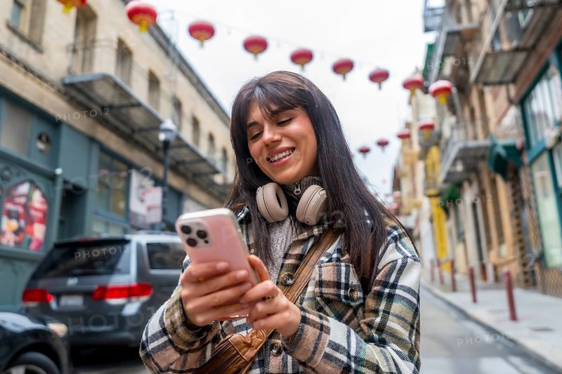 Young smiling woman exploring san francisco chinatown, browsing smartphone content and enjoying city life with headphones around her neck, reflecting urban travel and modern communication