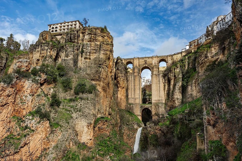 Wide angle view from the bottom of the gorge featuring the massive arches of Puente Nuevo bridge and a small waterfall in Ronda, Spain