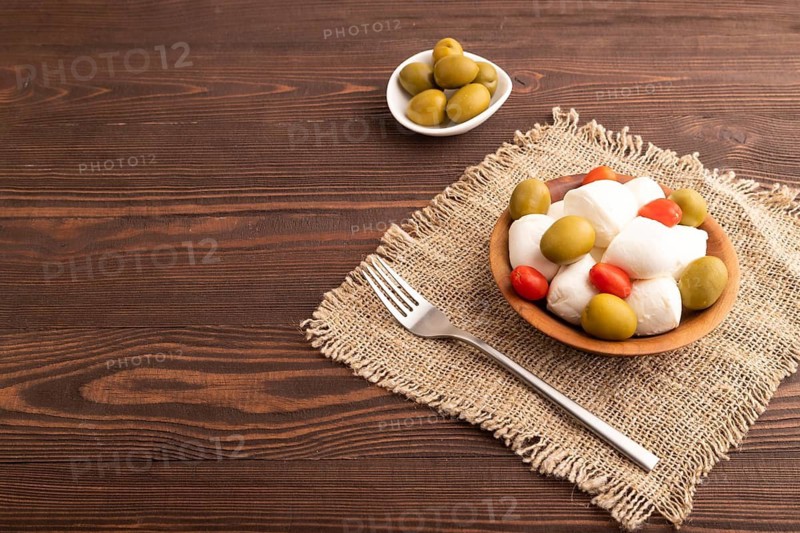 White Mozzarella cheese, with tomatoes and olives in wooden bowl on brown wooden background and linen textile, side view, copy space