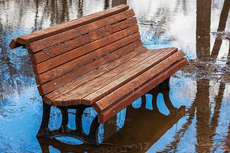 Empty brown stained long wooden sitting bench with black cast iron legs surrounded by flood water in public park flooded by heavy rainfall in spring, Ile des Moulins, Old Terrebonne, Quebec, Canada