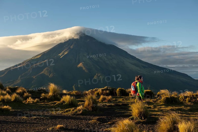A woman in hiking gear stands before the majestic Mount Taranaki in New Zealand, basking in the serene beauty of the volcanic landscape at sunrise