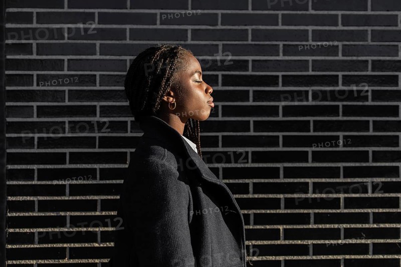 Side view of an African American businesswoman with braids, dressed in a professional coat, takes a peaceful mindful break against a brick wall backdrop, enjoying the sunlight with eyes closed