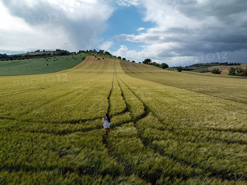 A young woman in a white dress walks through lush green fields in the Italian countryside. The landscape features vast, serene hills under a cloudy sky, embodying nature's tranquility