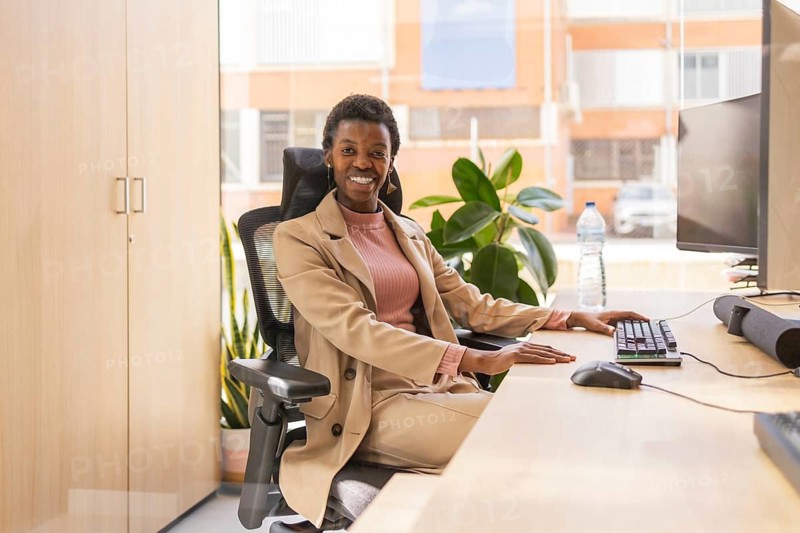A person wearing a suit sits at a desk, smiling confidently. The office is modern with natural light, plants, and a large computer monitor, creating a welcoming workspace