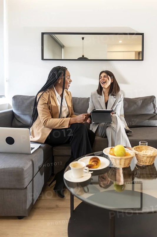 Two businesswomen are sitting on a sofa in a hotel room, laughing while working on a tablet during a business trip