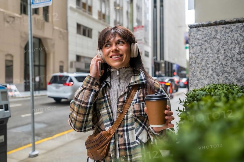 Woman holding a takeaway coffee cup and wearing headphones, engaging with her smartphone while walking and smiling on a bustling city street, embodying a cheerful urban lifestyle
