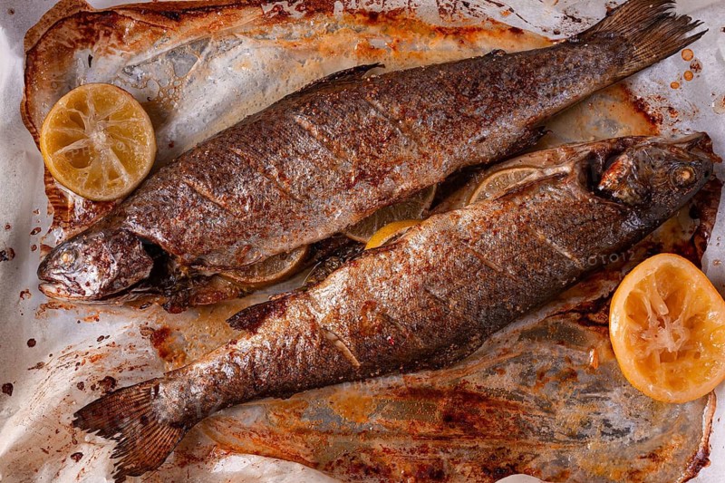 Two fish are being cooked on a baking sheet in a home kitchen. Lemon slices are placed beside the fish to add flavor. The dish shows grill marks and a brown color from baking