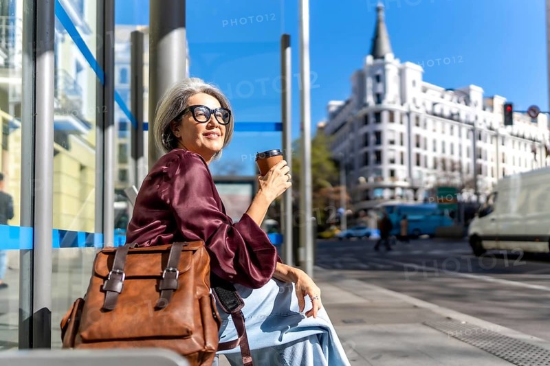 Senior woman is sitting at a city bus stop, holding a disposable coffee cup, embracing a relaxed urban lifestyle while commuting on a sunny morning in a european city
