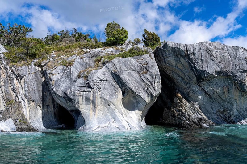 Marble caves (Cuevas de Mármol) with eroded rock formations on the turquoise waters of Lago General Carrera, overgrown rocky peak under a blue sky with clouds, Puerto Río Tranquilo, Aysén region, Chile