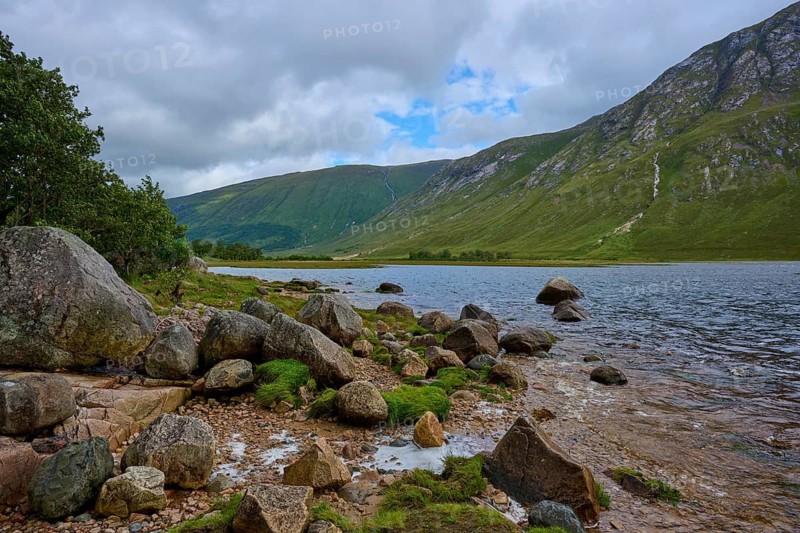 Rocks on the shore of a lake with green mountains in the background under cloudy sky, Loch Etive, Argyll and Bute, Scotland, United Kingdom, Great Britain