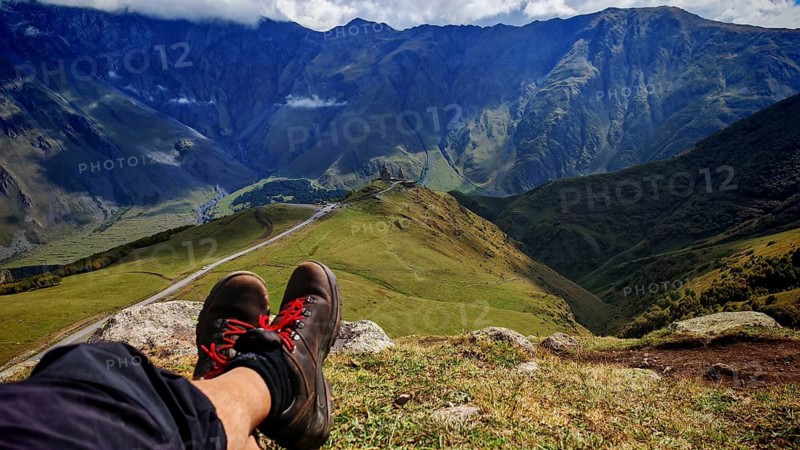 Perspective of a seated observer enjoying views of mountains, hiking boots with Gergeti church in the background, Stepantsminda, Caucasus, Georgia