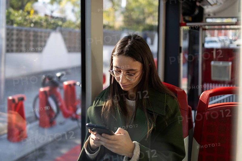 A young woman wearing glasses and a green coat looks at her smartphone while riding public transportation The dimly lit interior and window reflection add depth