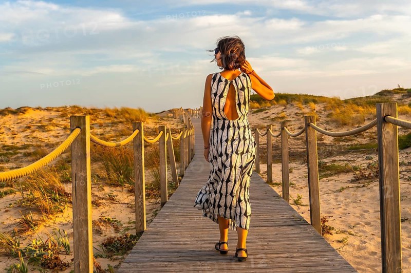 Woman walking along a wooden boardwalk through sand dunes towards the beach, enjoying the warm glow of the setting sun during her vacation in seignosse, france