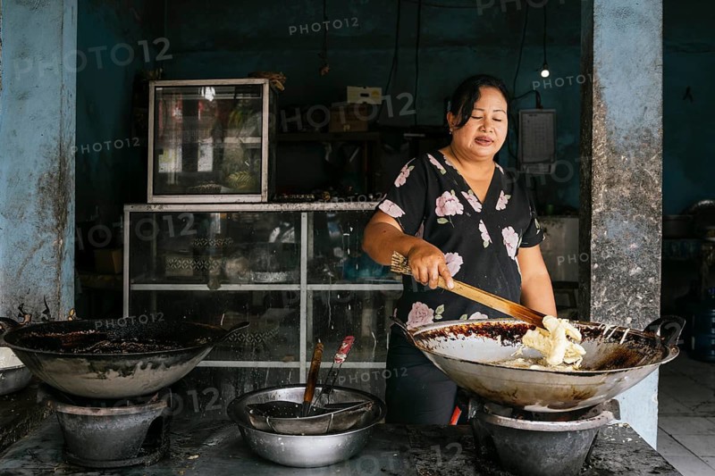 A woman prepares traditional street food in a market setting of Bali. She is frying snacks in a large wok, adding to the vibrant culinary culture of the region