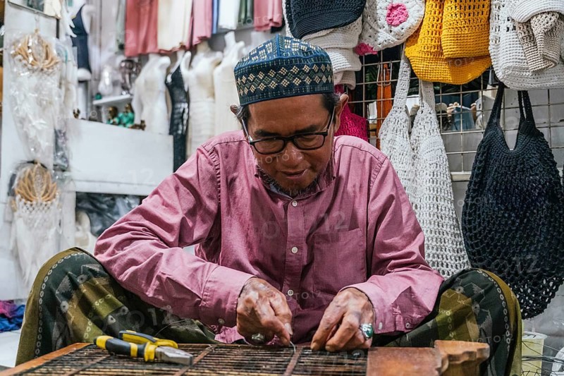 A balinese artisan wearing glasses focuses on crafting handmade goods in a vibrant local market. Colorful crochet bags adorn the background, reflecting craftsmanship