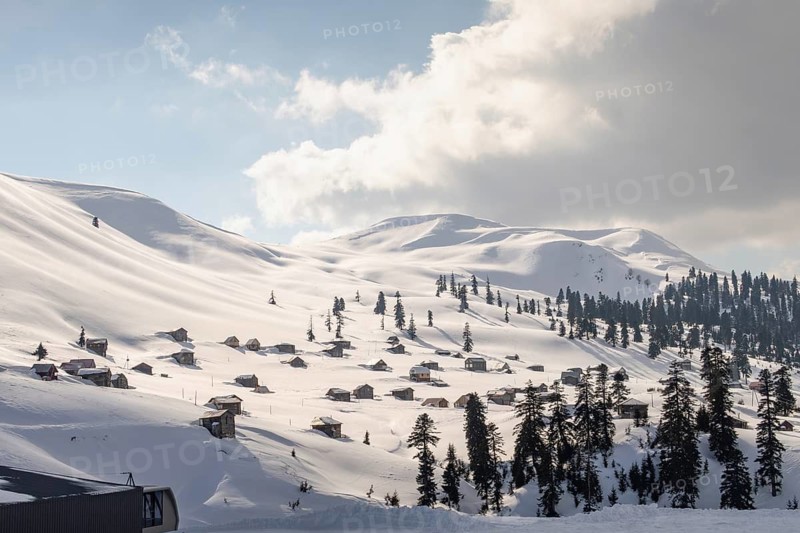 A stunning winter landscape in Georgia featuring snow-covered mountains dotted with quaint wooden cabins. Tall pine trees add contrast to the pure white snow under a partly cloudy sky