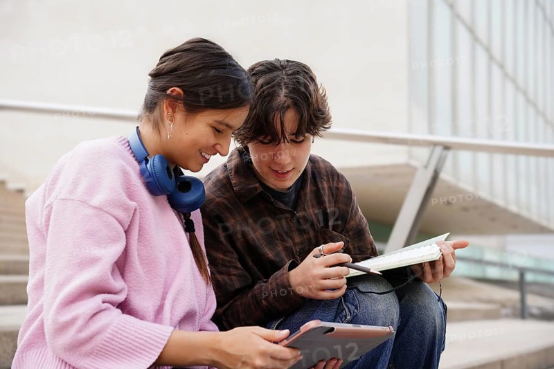 Two teenagers, a non-binary individual and a female, are studying together outdoors, using a tablet and notebook. The environment is casual, suggesting a scene of young educational collaboration