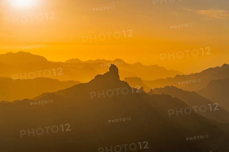 A captivating view of Roque Nublo under a golden sunset, showcasing layers of mountains bathed in warm light, casting dramatic silhouettes across the rugged landscape of Gran Canaria